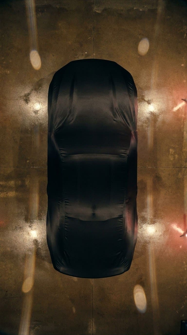 An overhead view of a car under a fitted black satin cover on a garage floor, warm lights glowing on the concrete around it.