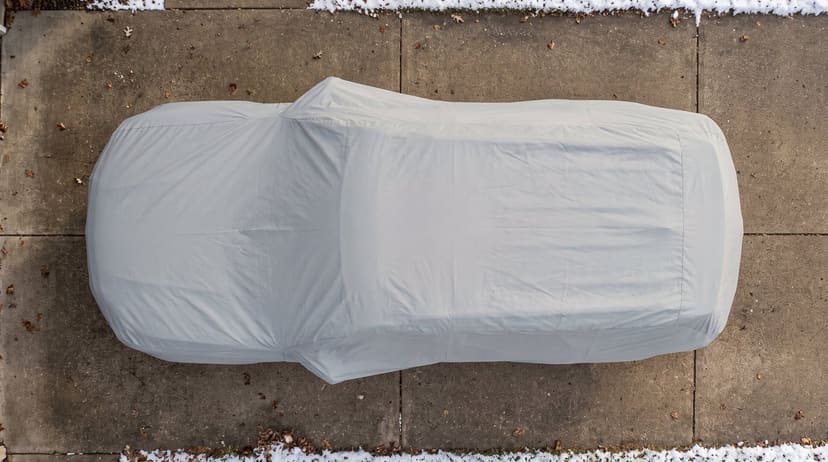 Top-down view of a light grey cover fitted over an SUV on a concrete driveway with fallen leaves and a little snow at the edges.