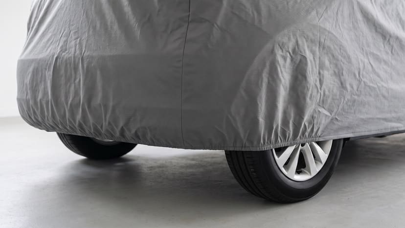 Close up of the rear corner of a car under a medium grey matte cover, its elastic hem gathered above the tires on a pale garage floor.