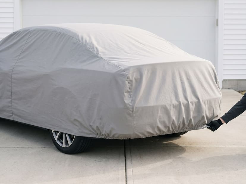 A light grey cover on a sedan in a driveway, seen from the rear corner, a gloved hand drawing the elastic rear hem in under the bumper