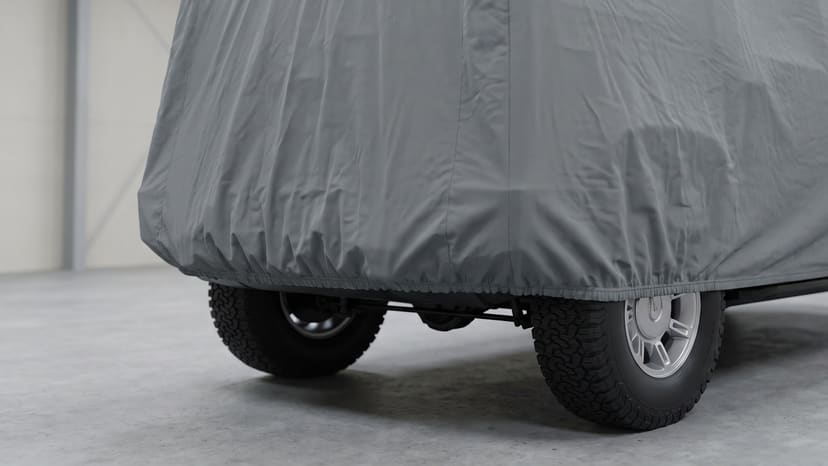 Close up of the rear of a boxy off-road SUV under a medium grey matte cover, its elastic hem gathered above the all-terrain tires on a concrete floor.