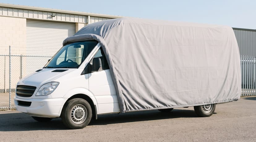 A white high-roof van with a light grey cover drawn back off the front, the door mirror in the open, on a business lot in front of a roller-door unit and a chain-link fence.