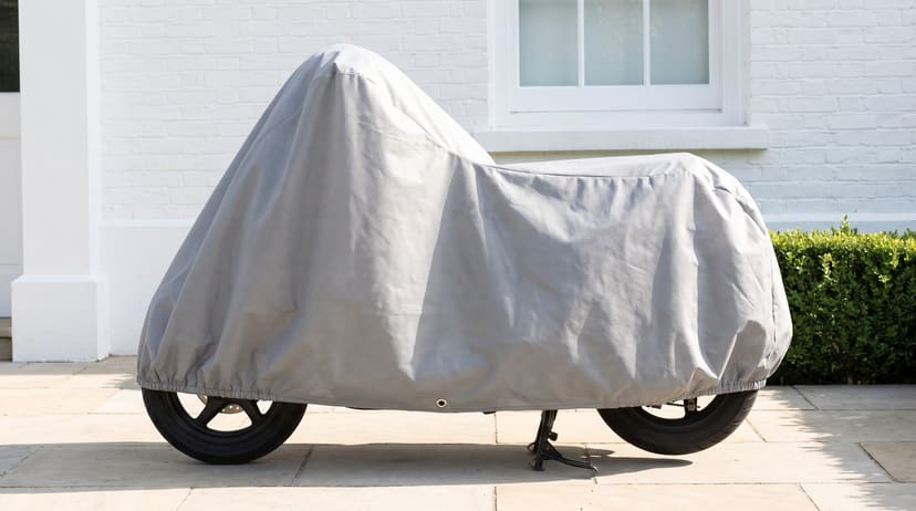 A scooter under a light grey cover on pale paving by a white house.