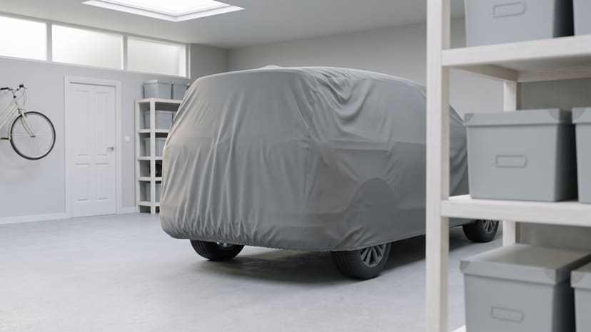The rear of a van under a medium grey matte cover in a bright home garage, seen past shelves of storage boxes, a bicycle on the far wall.
