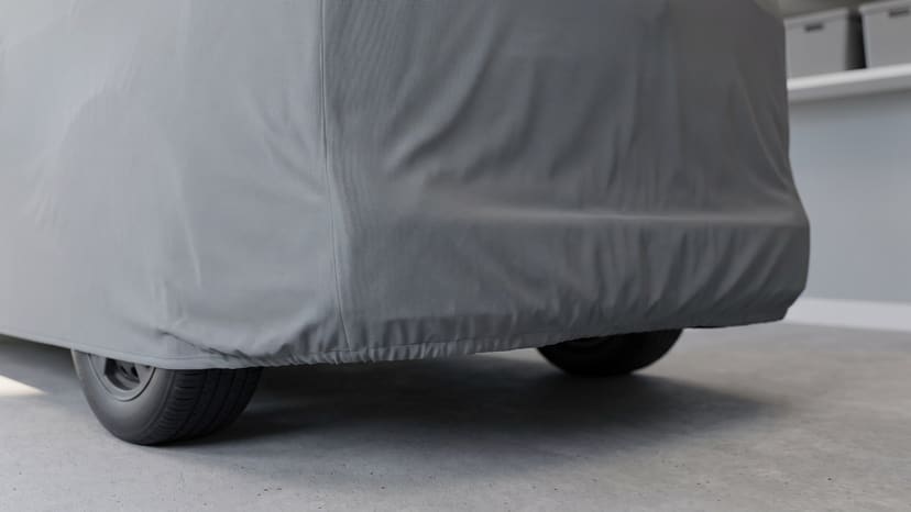 Close up of the rear corner of a van under a medium grey matte cover, its elastic hem gathered above the tires on a concrete garage floor.