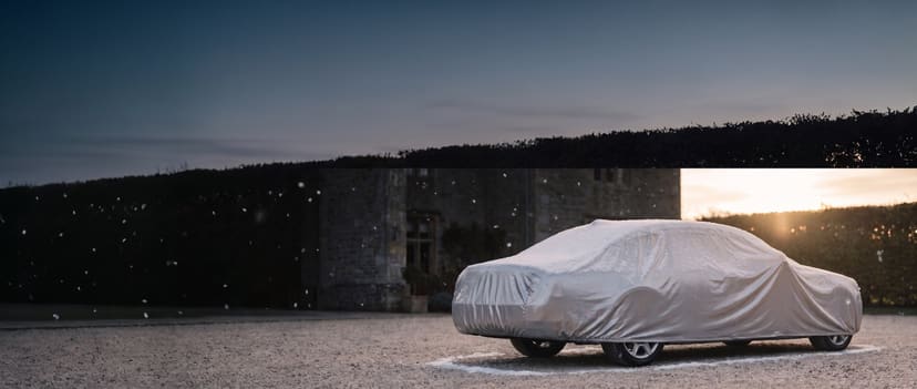 Silver cover on a full-size sedan with a light dusting of snow beside a stone house at dusk