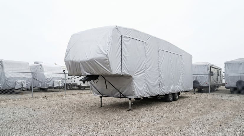 A fifth-wheel trailer on its landing gear in an RV storage lot, under a fitted silver cover.