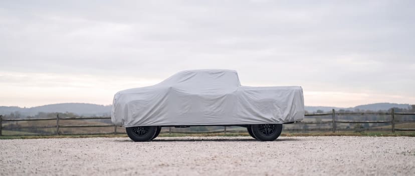 A full-size pickup under a silver outdoor cover on a gravel ranch drive, a fence and hills behind.