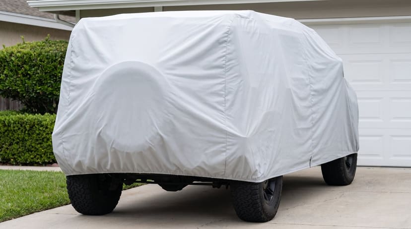 The rear of a big boxy off-road SUV under a light grey cover, its spare wheel a round swell, on a concrete driveway beside a white garage door.