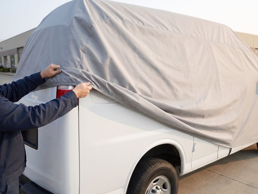 Hands rolling a light grey cover forward off the back of a white high-roof van on a business lot