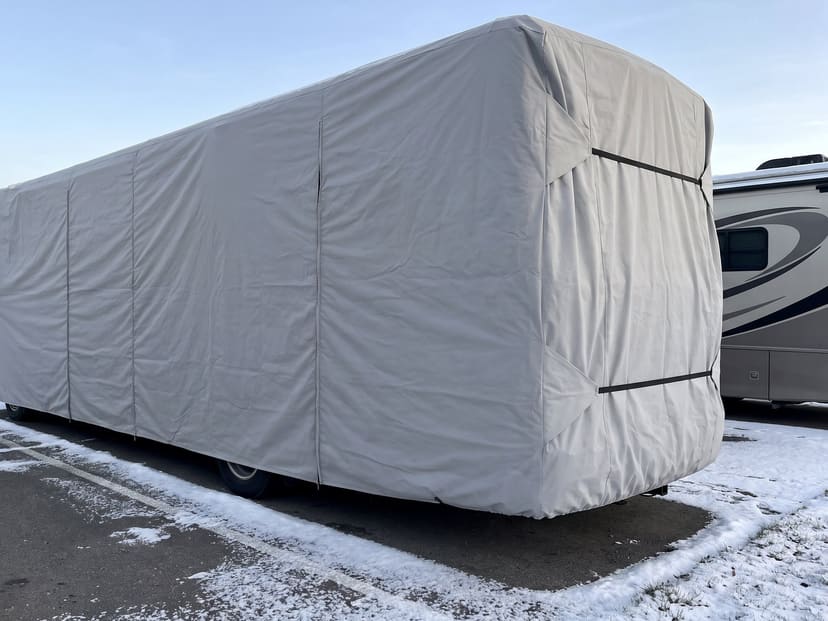 A Class A motorhome in a snowy storage lot, under a fitted light grey cover strapped at the rear.
