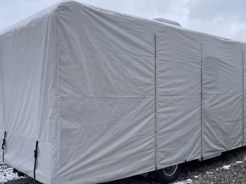 The rear of a motorhome in a snowy storage lot, under a fitted light grey cover.