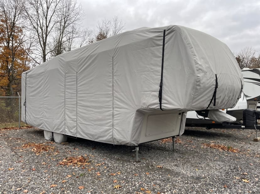 A fifth-wheel trailer on its landing gear in an autumn storage lot, under a fitted light grey cover.