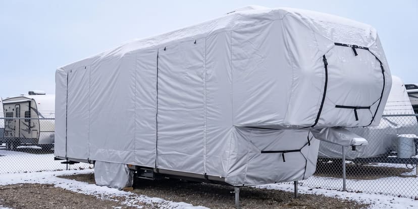 A fifth-wheel trailer in a snowy RV storage lot, under a fitted silver cover.