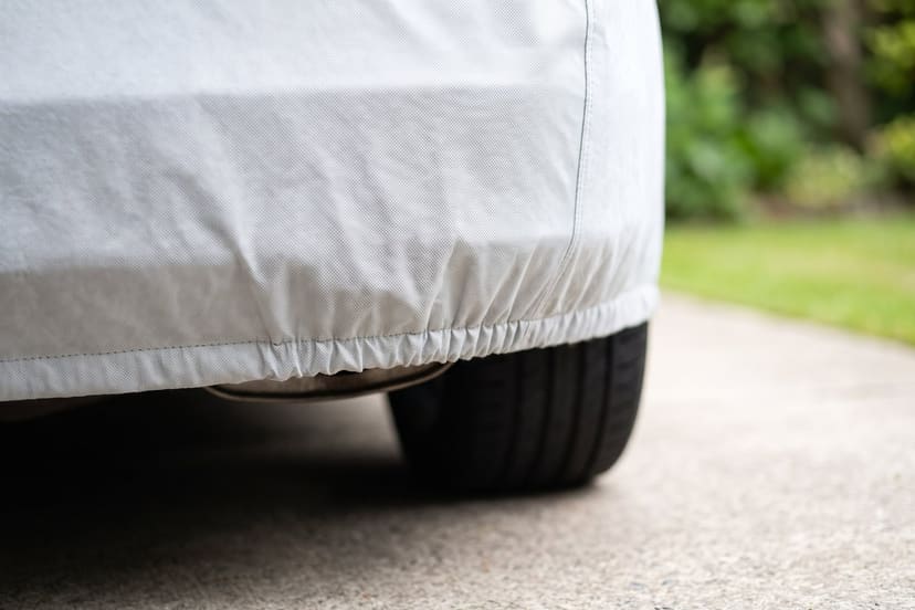 The elastic hem of a light grey cover gathered under the rear bumper above a tyre on a concrete driveway
