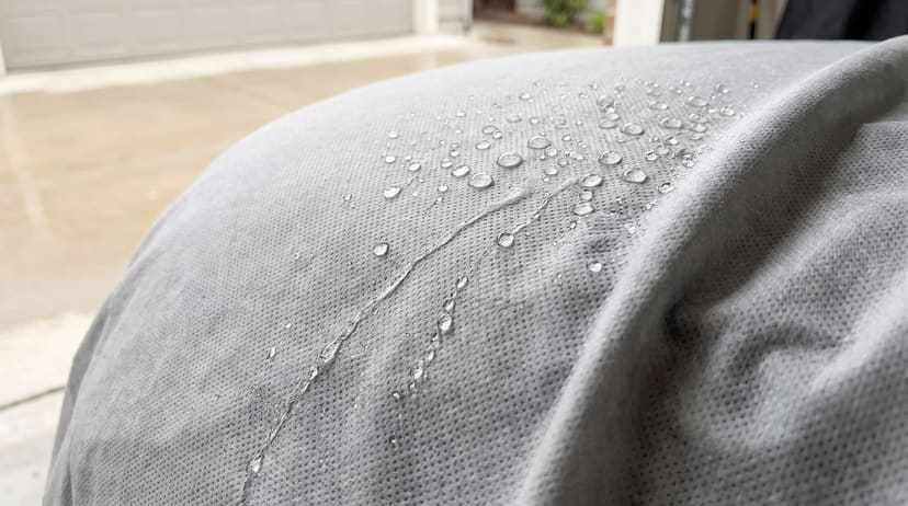 Close-up of rain beading on a grey non-woven cover fabric across the curve of a covered car
