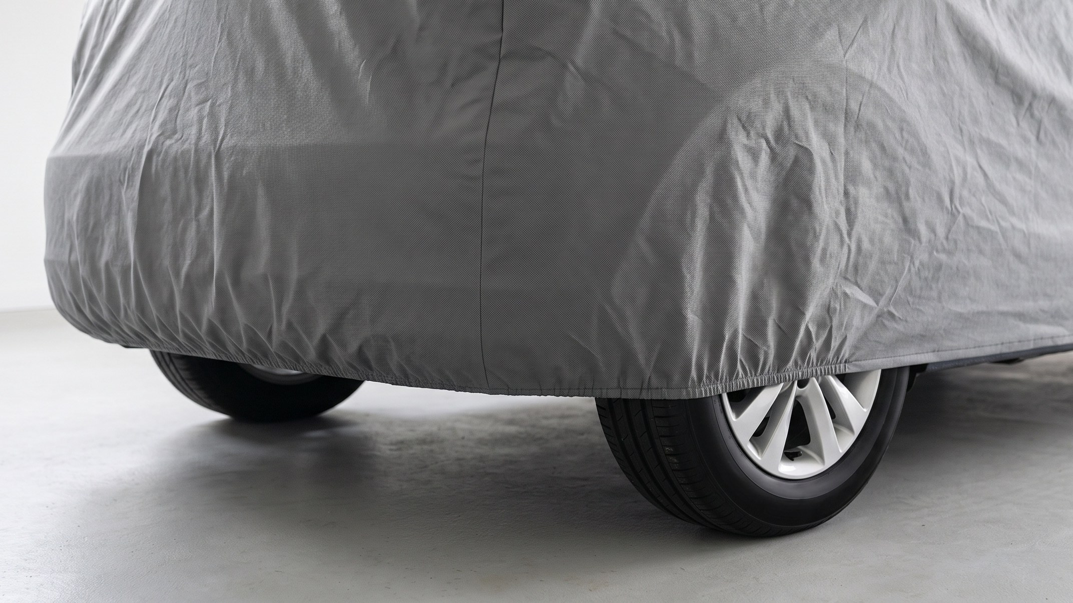 The rear corner of a car under a grey cover, its elastic hem drawn in under the bumper above the rear wheels