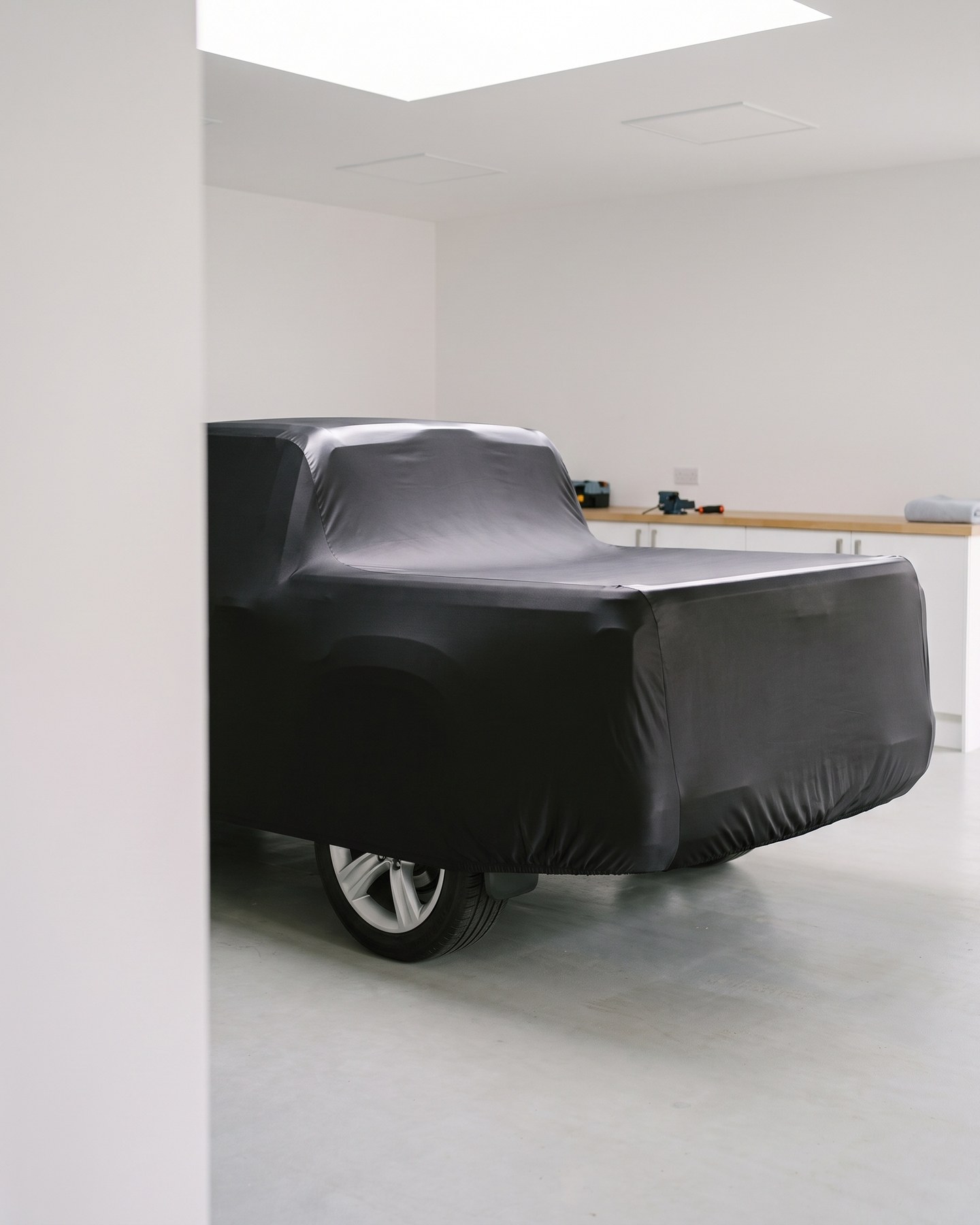 The rear of a full-size pickup under a black stretch indoor cover in a bright garage