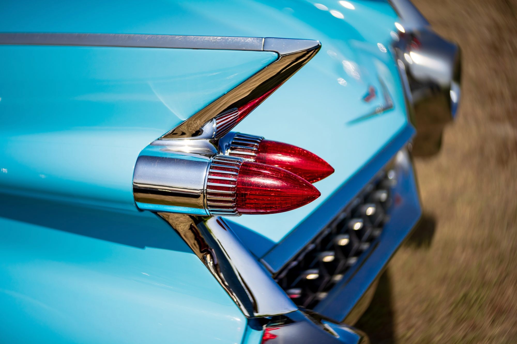 A close-up of a chrome tailfin and twin red bullet-shaped taillights on a pale turquoise vintage American car, sunlit, with a blurred tan landscape behind it.