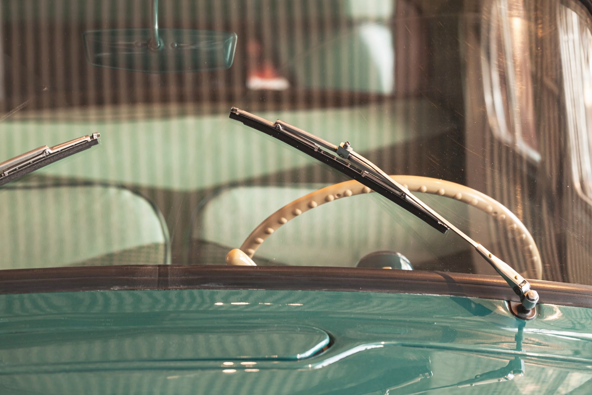 Chrome wiper arms rest on the windshield of a vintage teal car, seen from outside through the glass toward a cream steering wheel with a knobbed rim and mint-green seats.
