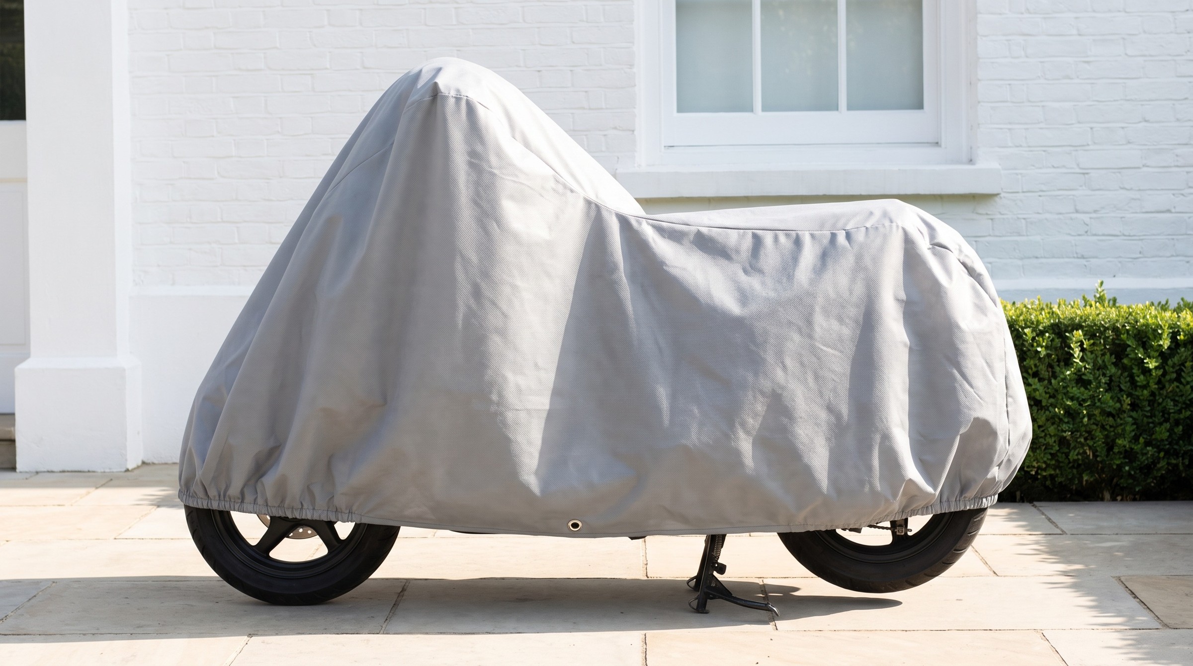 A scooter under a light grey cover on pale paving by a white house.