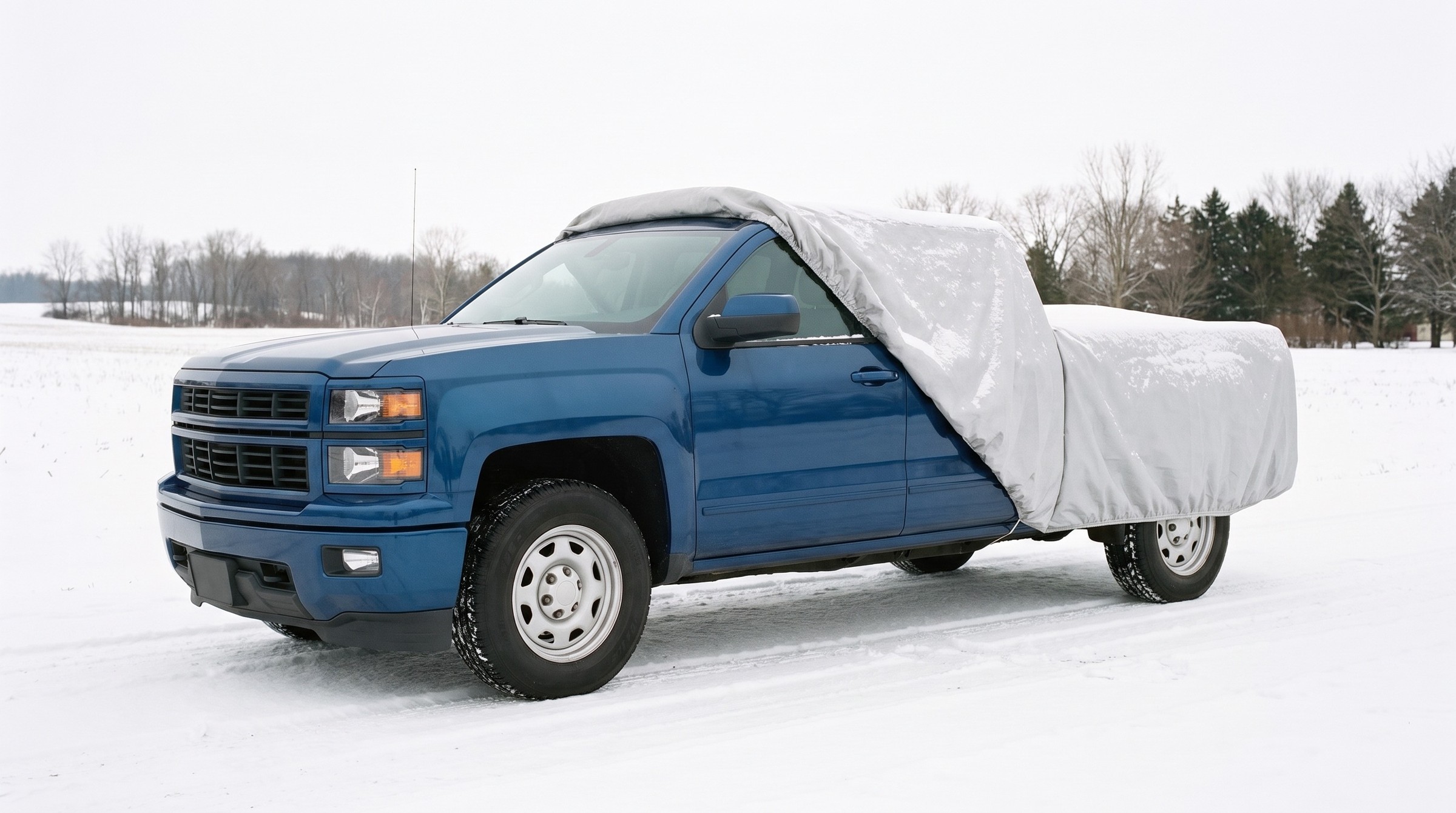 A blue pickup with a silver cover drawn back off the front, snow on the cover, the door mirror in the open, on a snowy track in an open field.