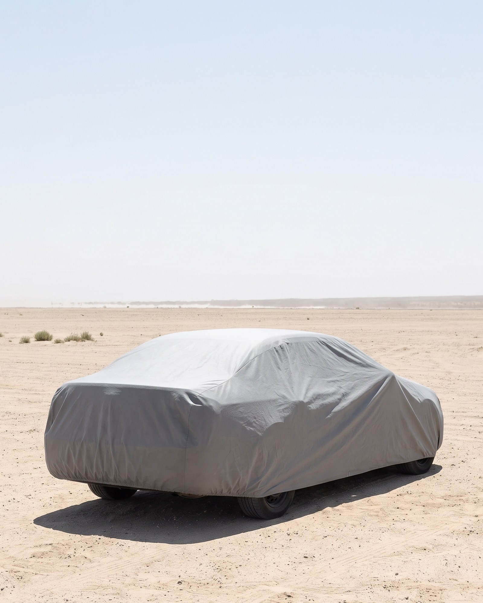 A sedan under a grey cover, seen from the rear three-quarter on open desert sand under a pale, hazy sky in hard sun