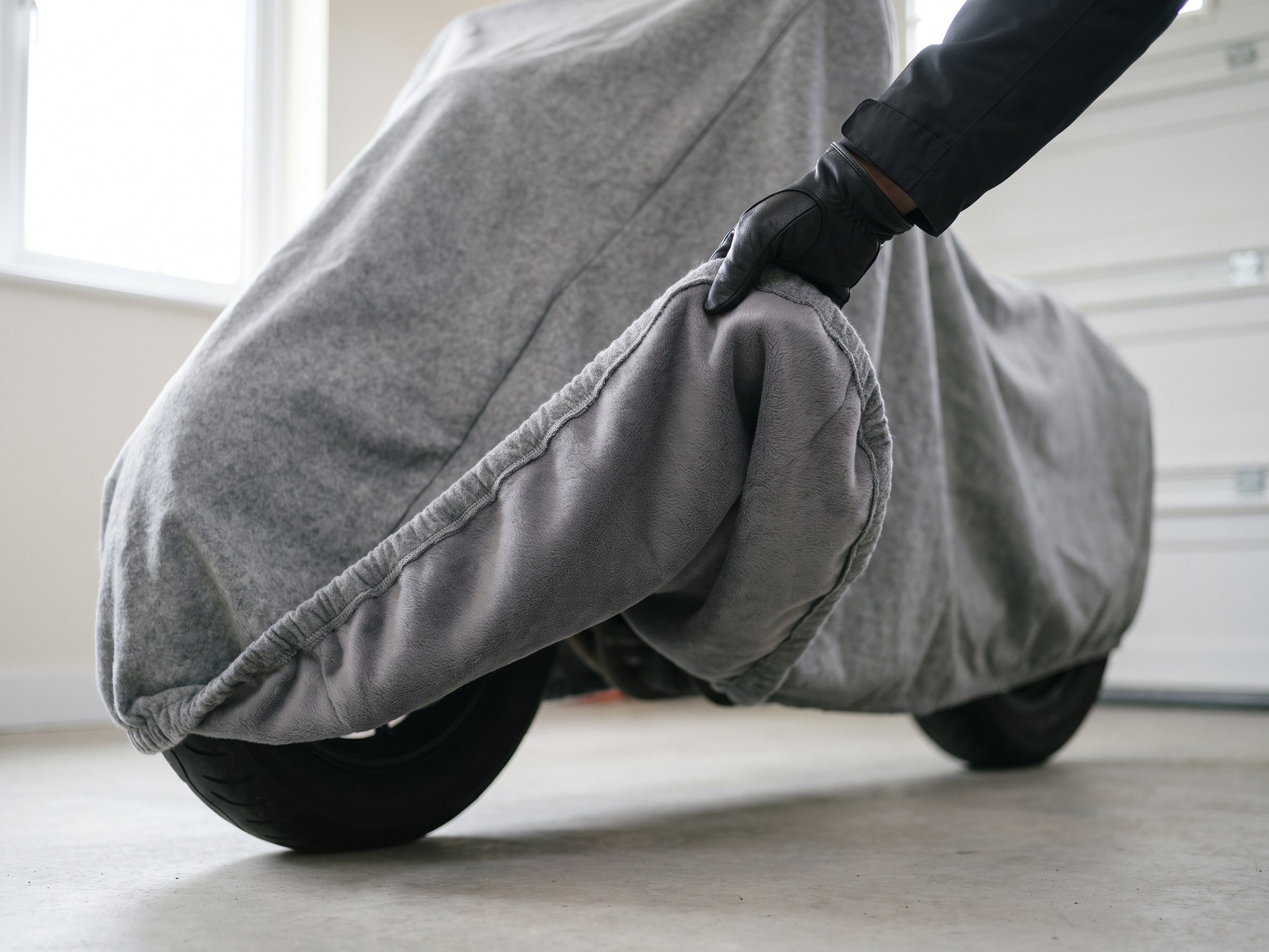 A gloved hand turning back the elastic hem of a gray motorcycle cover over the front wheel to show the soft gray fleece lining inside, in a bright garage.