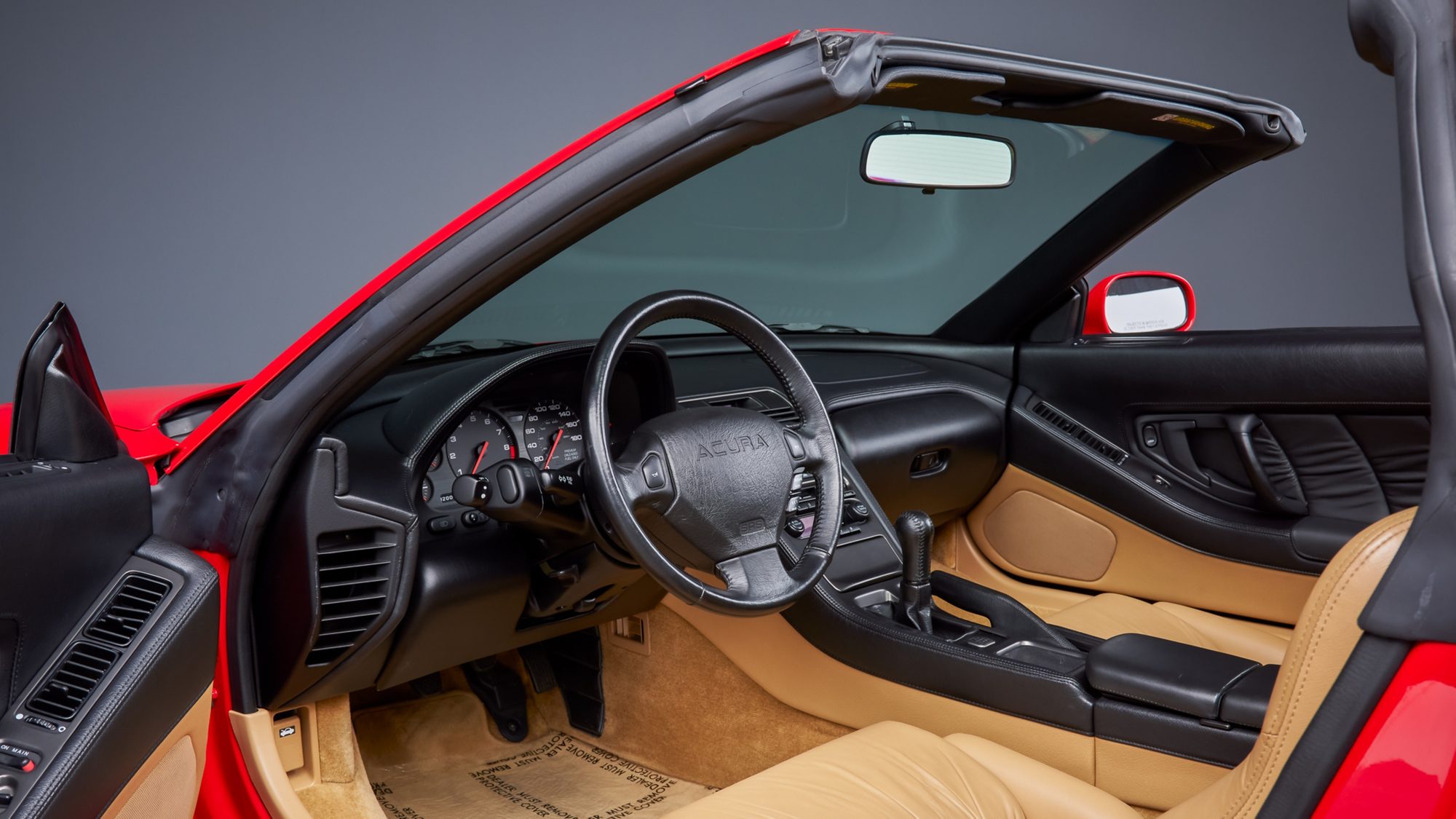 The NSX's black dashboard and tan leather cabin seen through the open driver's door with the targa roof panel off: a leather-wrapped steering wheel, analog gauges and a manual shifter.