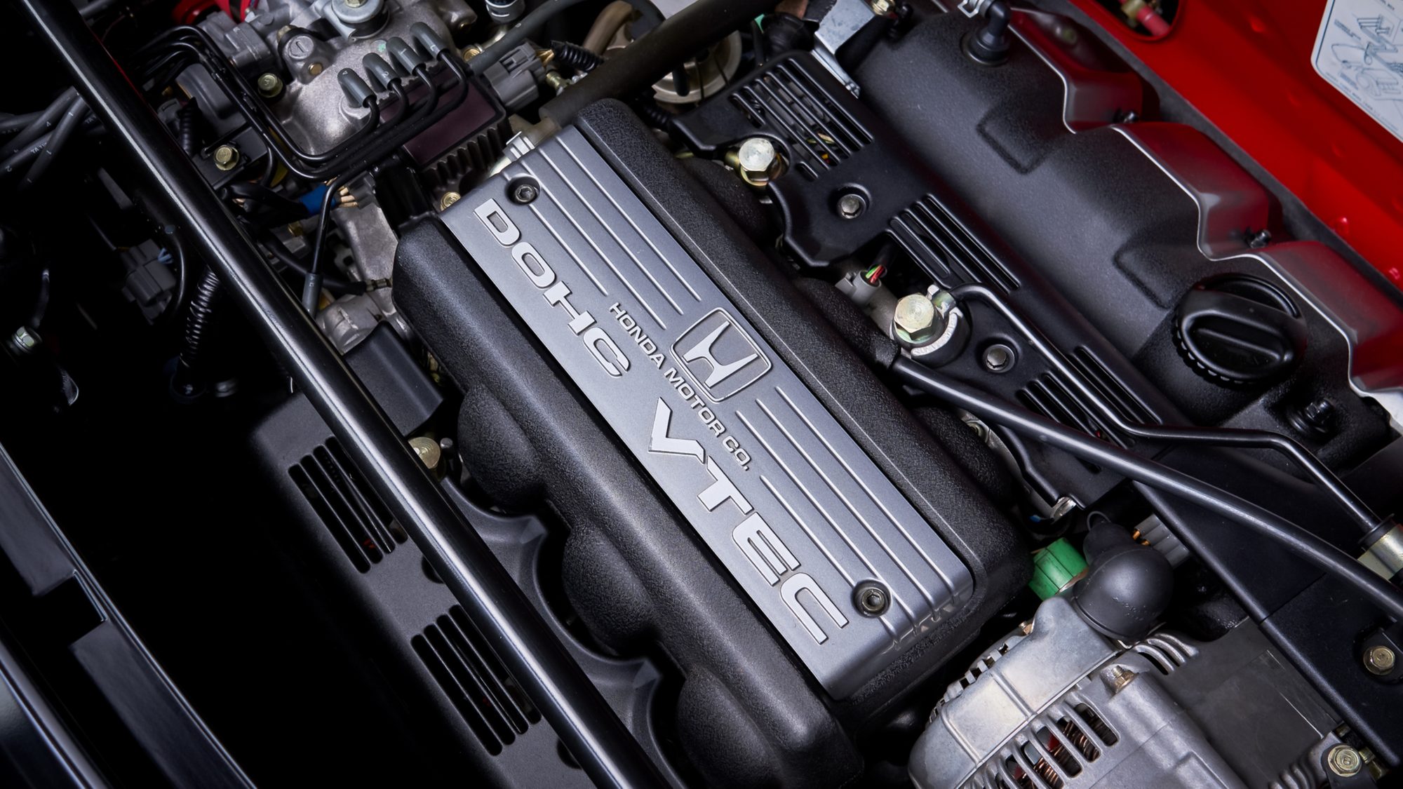 The NSX's V6 seen from above in its engine bay, a ribbed silver cover marked DOHC VTEC and Honda Motor Co. with the Honda H badge, surrounded by hoses and wiring.
