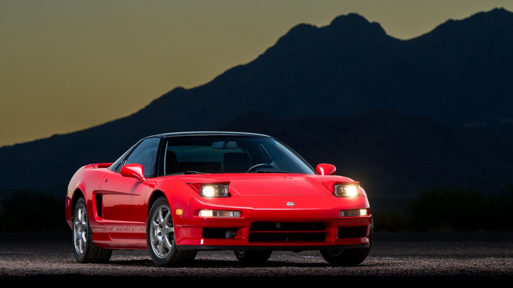 A red first-generation Acura NSX in front three-quarter view at dusk, its pop-up headlights raised and lit, against a dark mountain silhouette.