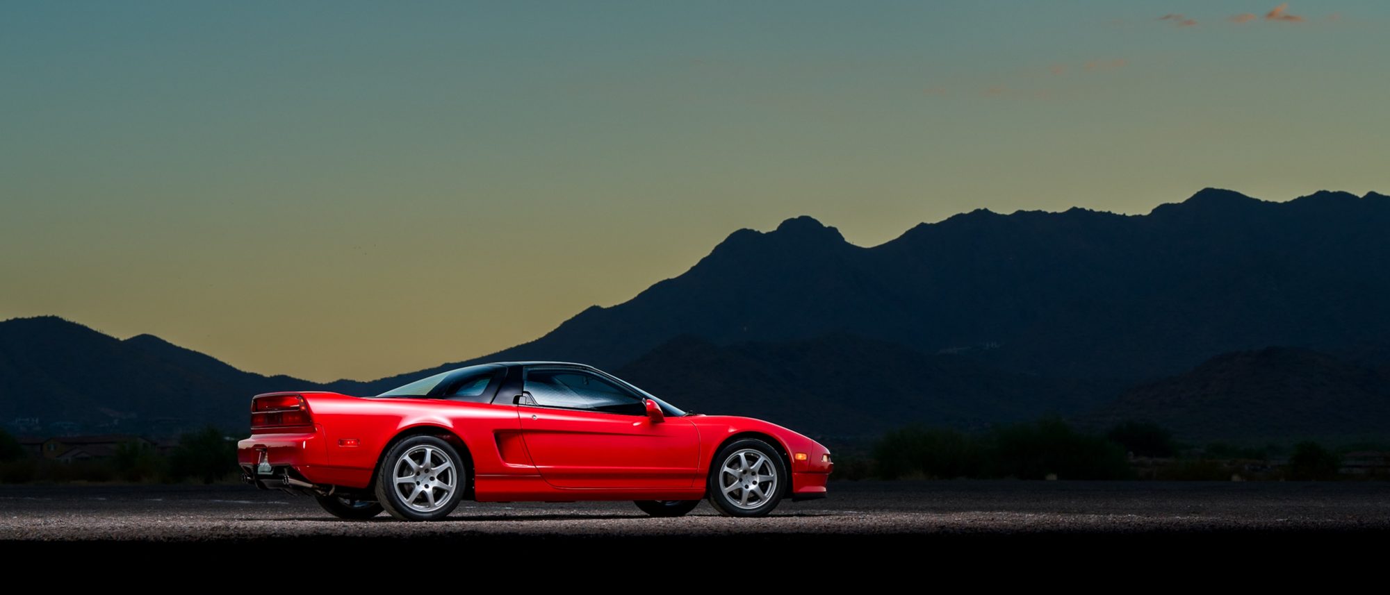 A red first-generation Acura NSX seen from the side at dusk, its low, wedge-shaped silhouette set against a mountain skyline.