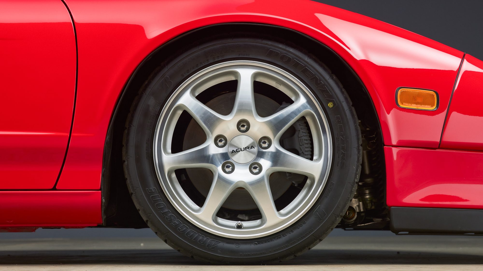 Close-up of a red NSX's seven-spoke alloy wheel with an Acura center cap and a Bridgestone Potenza tire, an amber side marker on the front fender beside it.