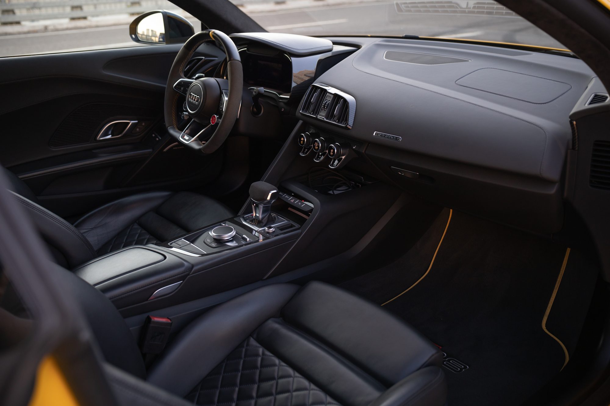 The cabin of a second-generation Audi R8 seen through the passenger door: black quilted leather seats, a suede-trimmed steering wheel, the digital instrument display and yellow-trimmed floor mats