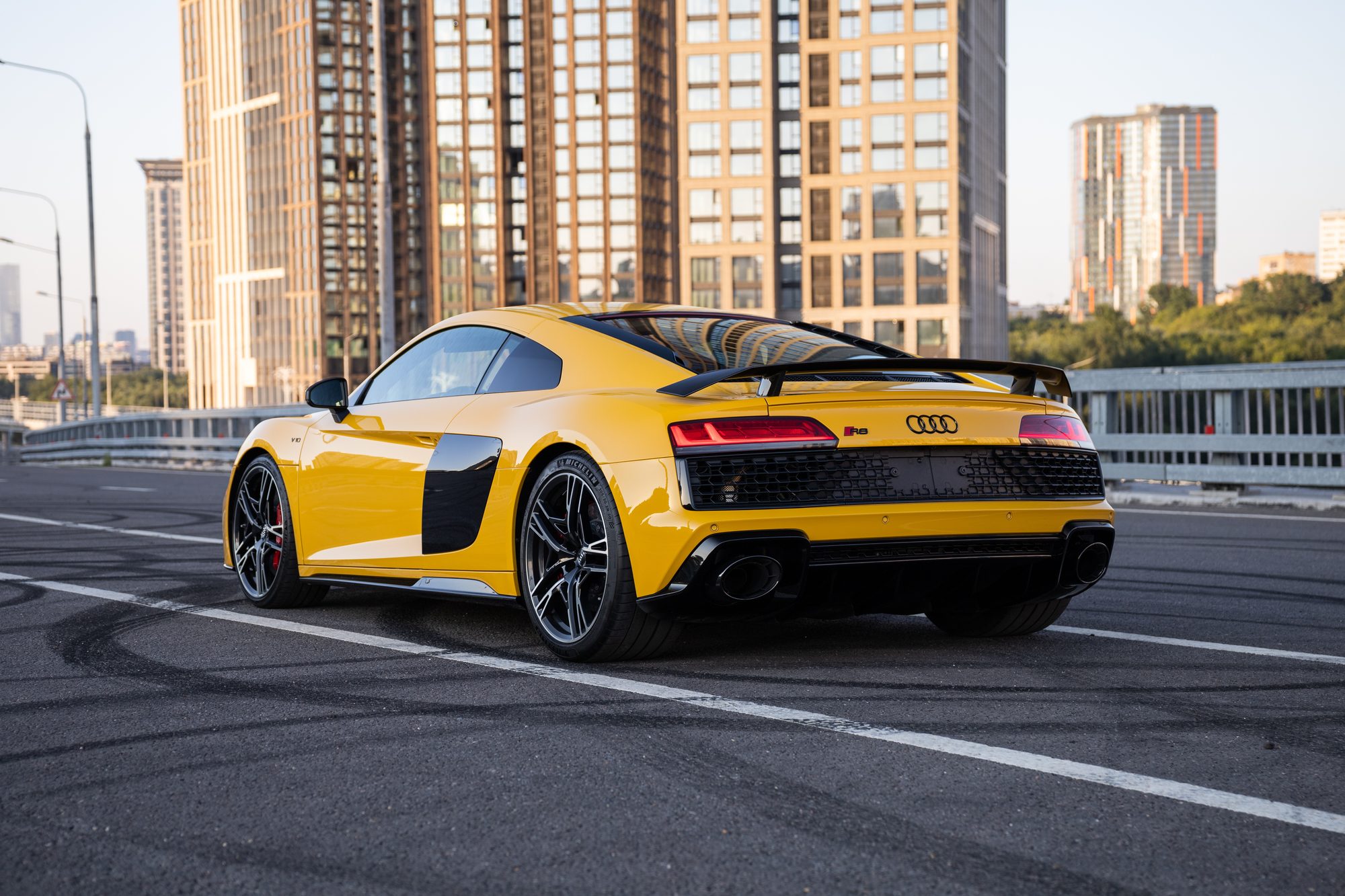 A yellow second-generation Audi R8 coupe seen from the rear three-quarter angle on an elevated city road, with a fixed rear wing, twin oval tailpipes and high-rise towers behind it