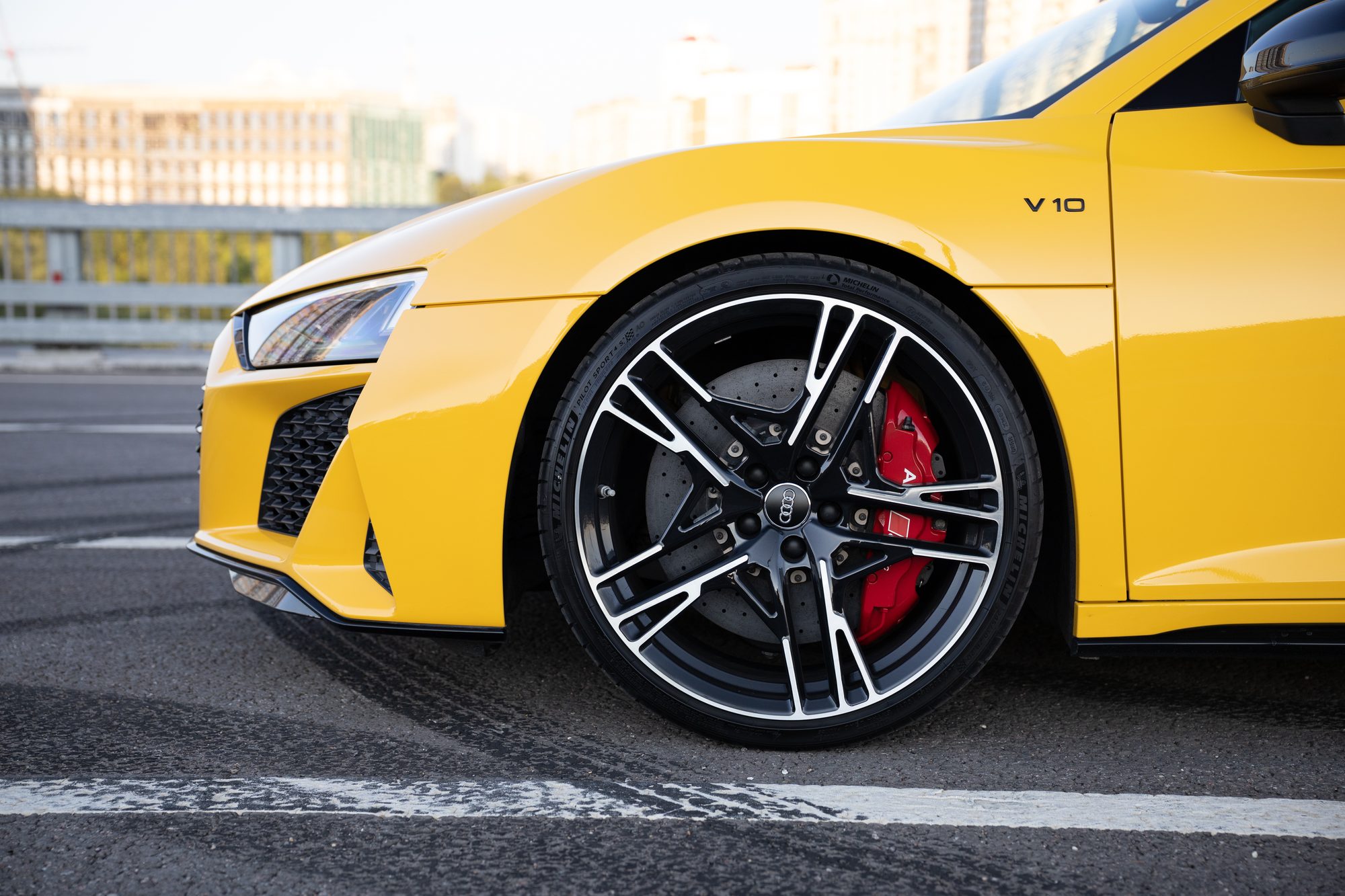 The front wheel and fender of a yellow second-generation Audi R8, with a V10 badge on the fender and a red brake caliper and drilled disc behind a black two-tone wheel