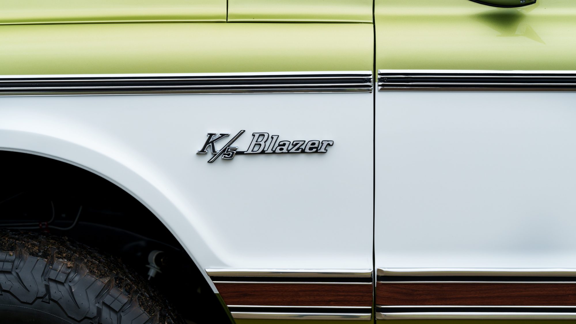 A close-up of the chrome K/5 Blazer script on the white front fender of a restored Chevrolet Blazer, above a wood-grain side stripe