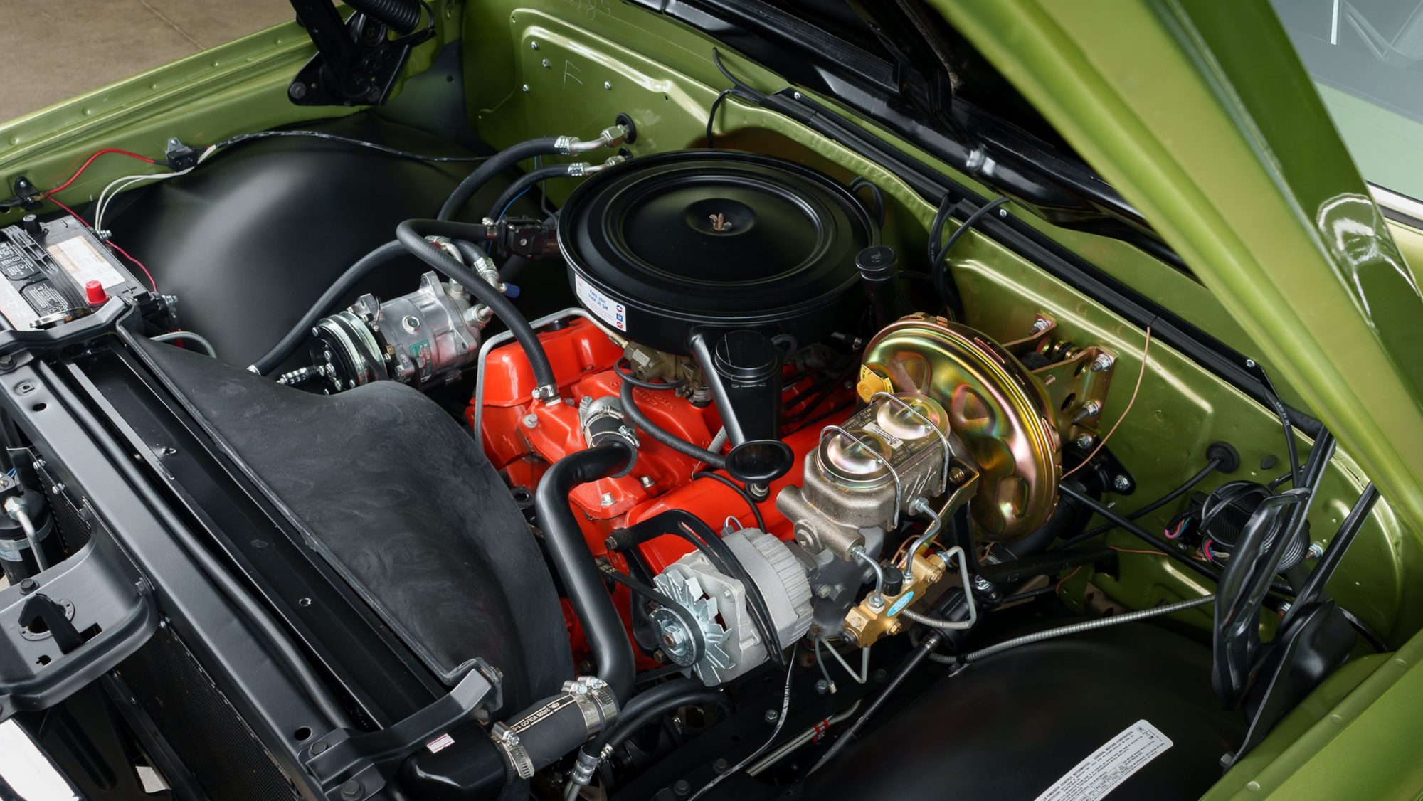 The engine bay of a restored Chevrolet K5 Blazer: an orange-painted V8 under a black air cleaner, with an air conditioning compressor at left and a gold-finished brake booster with its master cylinder at right