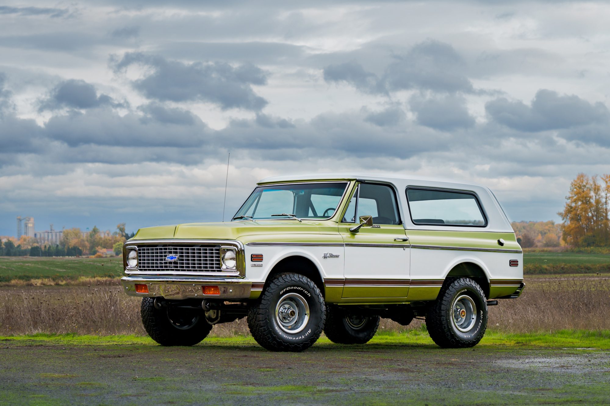 A restored early-1970s Chevrolet K5 Blazer in green and white with a white hardtop and a wood-grain side stripe, on large off-road tires, parked in a field under a cloudy sky, seen from the front three-quarter angle