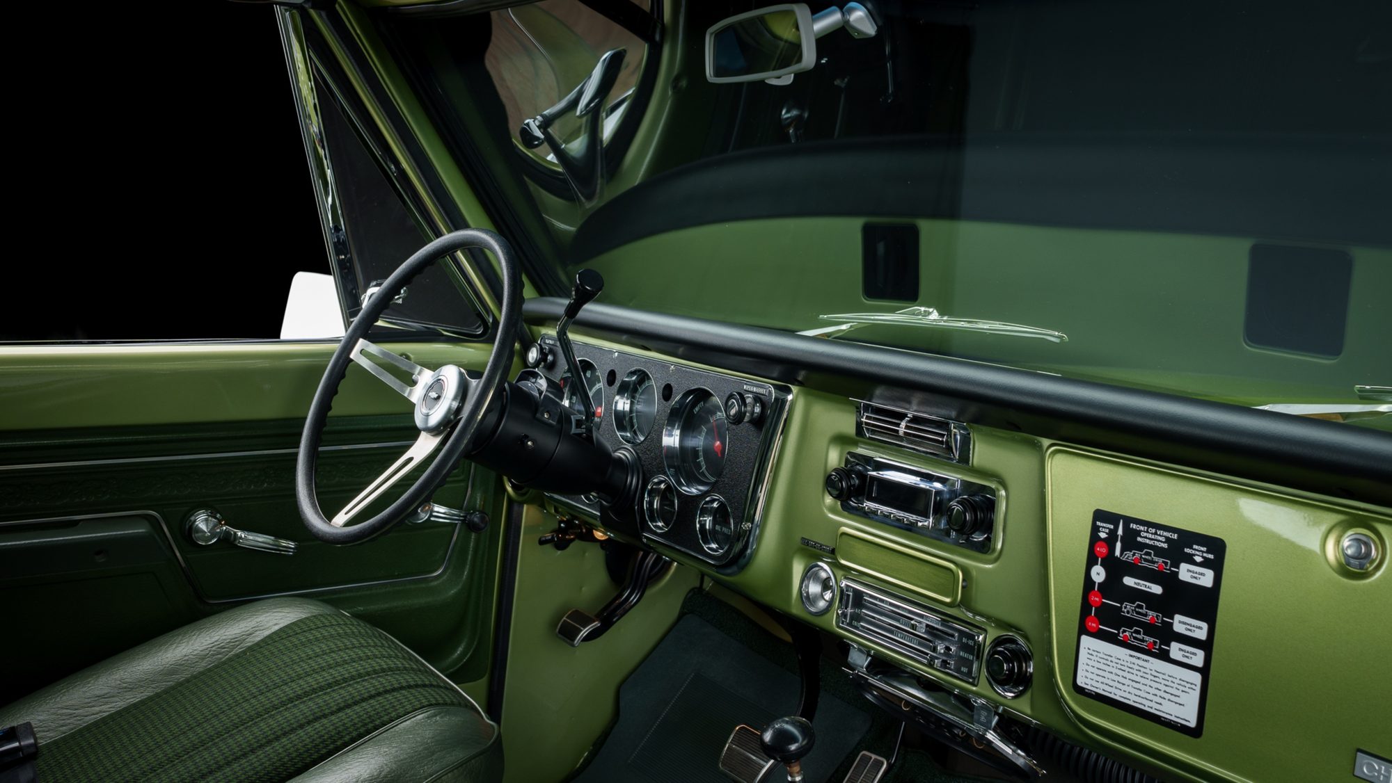 The green interior of a restored Chevrolet K5 Blazer: a slotted-spoke steering wheel, round gauges in a black instrument panel, a chrome radio and heater controls in the center of the dash, and a four-wheel-drive operating instructions decal on the glovebox door