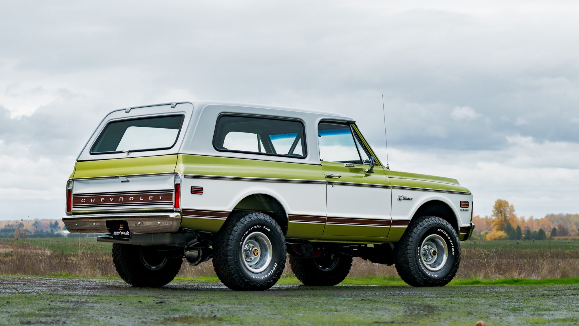 A restored early-1970s Chevrolet K5 Blazer in green and white, seen from the rear three-quarter angle in a field, with CHEVROLET lettering across a wood-grain tailgate panel and large off-road tires