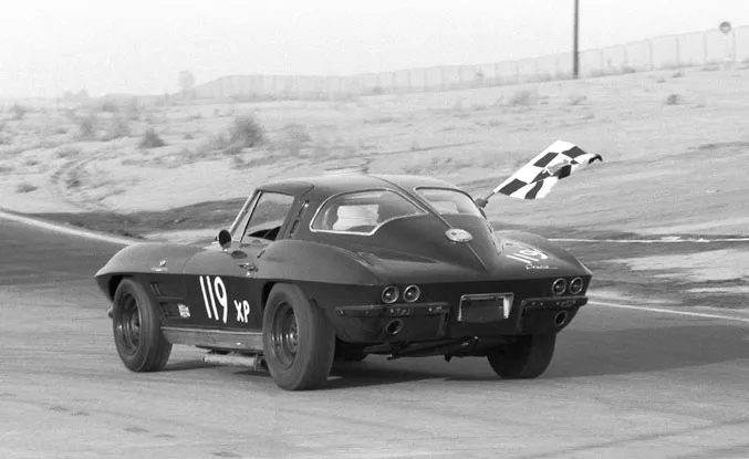 A black and white photo of a 1963 Corvette Sting Ray split-window race car, number 119 XP, on track with a checkered flag flying from the passenger window