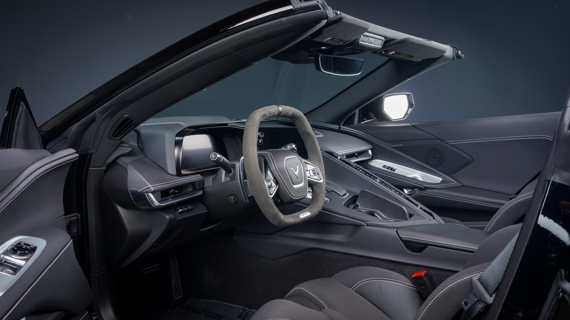The Corvette Z06's black leather and suede cockpit, seen through the open driver's door with the convertible top lowered and the low, wide dashboard visible