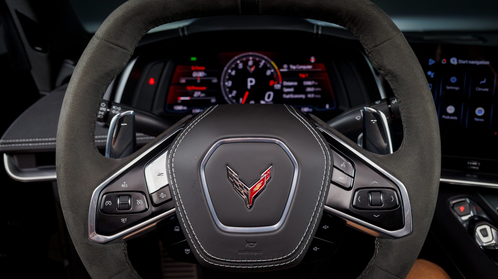A close view through the Z06's suede steering wheel showing the digital instrument cluster, the crossed-flags Corvette badge on the airbag cover, and wheel-mounted paddle shifters