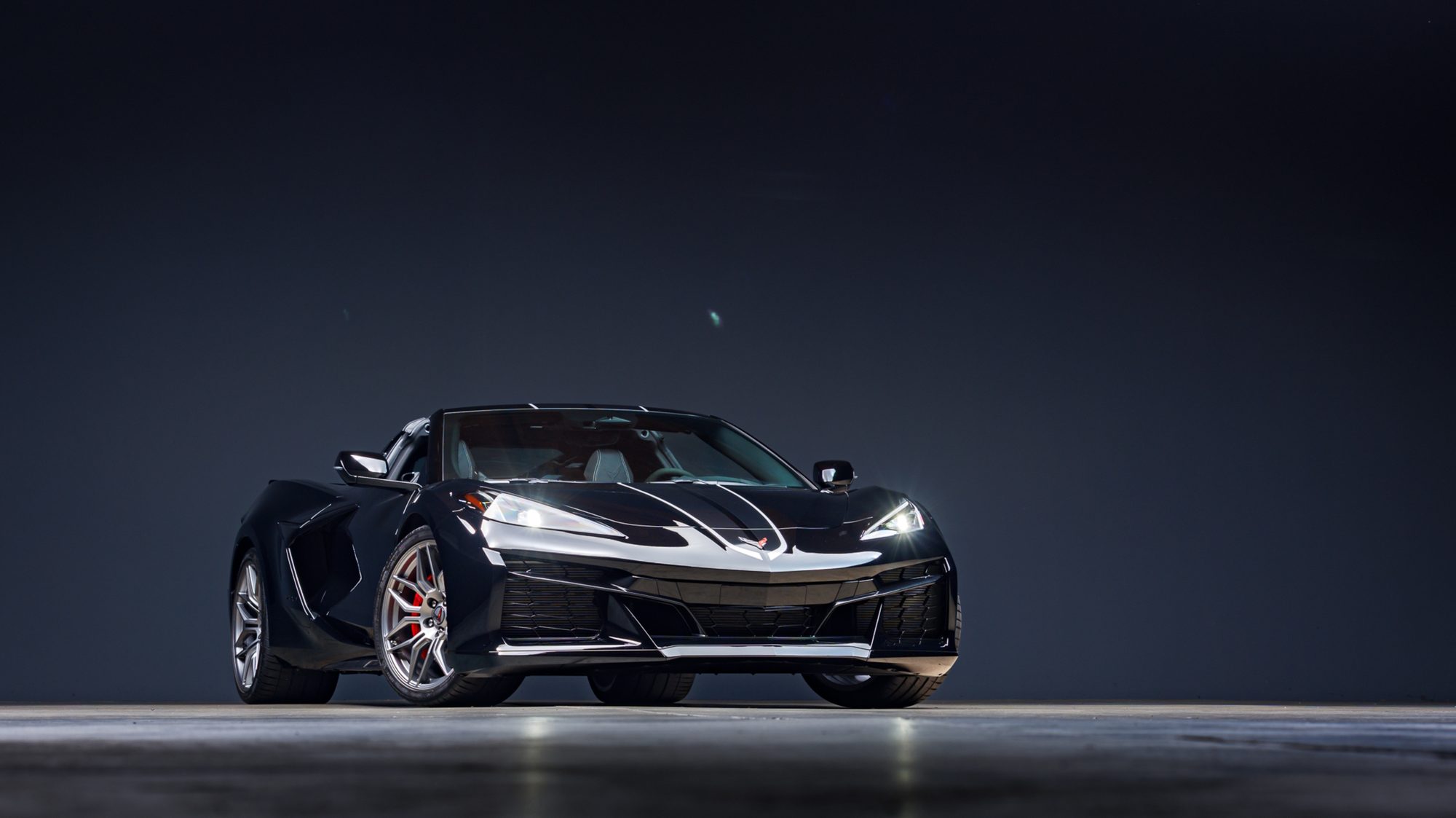 A black Chevrolet Corvette Z06 convertible seen from the front three-quarter angle in a dark studio, headlights lit, showing its wide front splitter and low stance
