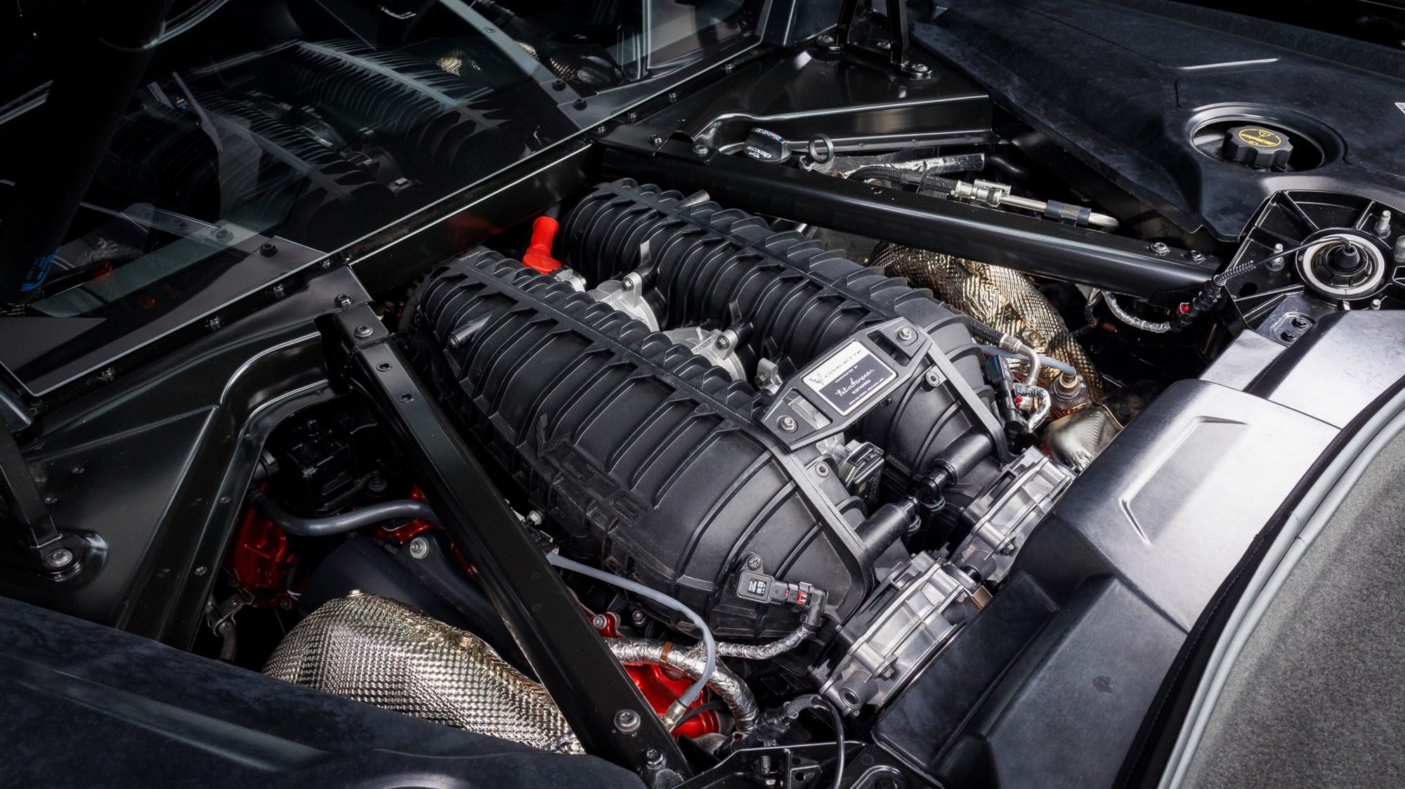 Close-up of the LT6 V8 engine mounted behind the seats in a Corvette Z06, showing its ribbed intake manifold and the builder's signed plaque on top, with the dry-sump oil tank's fill cap and loop dipstick beyond it