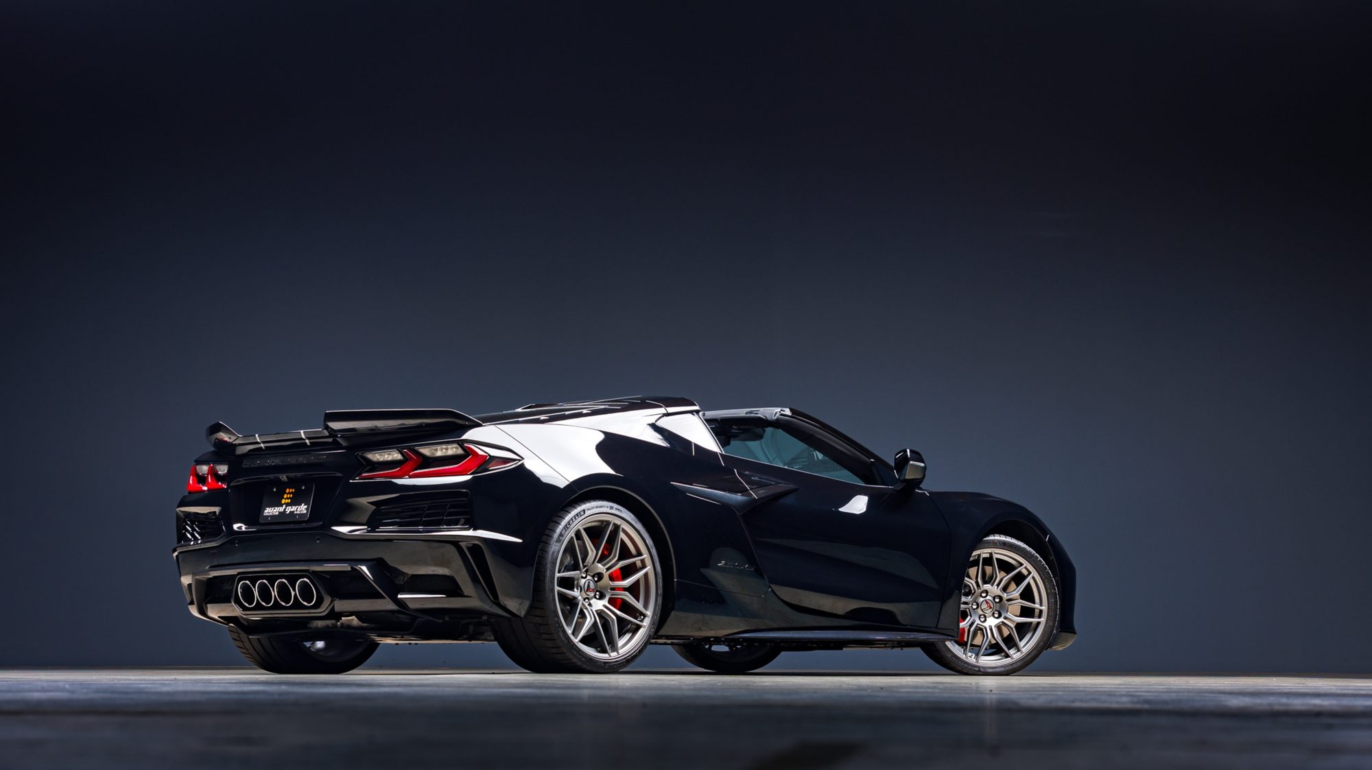 A black Corvette Z06 convertible seen from the rear three-quarter angle in a studio, its quad exhaust tips, rear spoiler and wide diffuser visible