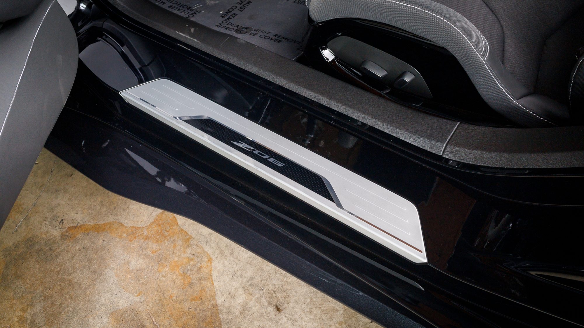 A brushed metal door sill plate on a Corvette Z06 engraved with the Z06 name, next to the grey seat bolster and a strip of carpet