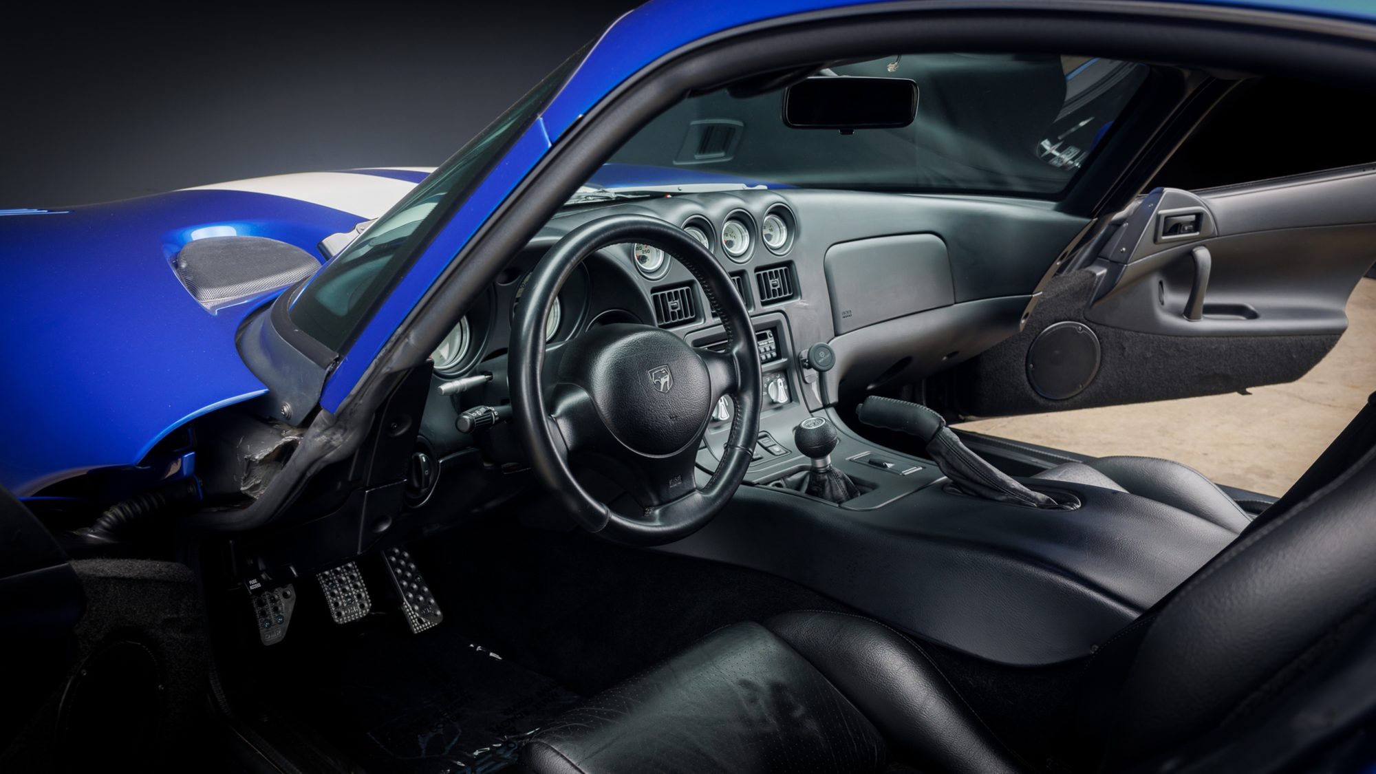 The Dodge Viper's cockpit seen through the open driver's door, showing a black leather seat, round gauges, the steering wheel, a manual shifter and drilled metal pedals.