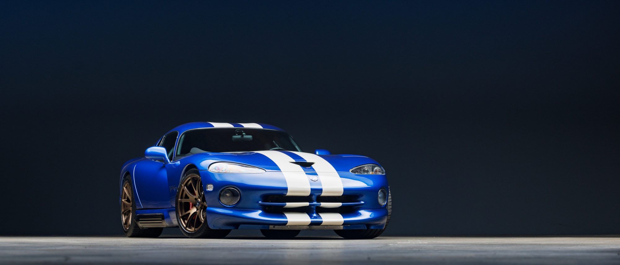 A blue Dodge Viper GTS coupe with white racing stripes, photographed from the front three-quarter angle against a dark studio backdrop, with bronze wheels and red brake calipers.
